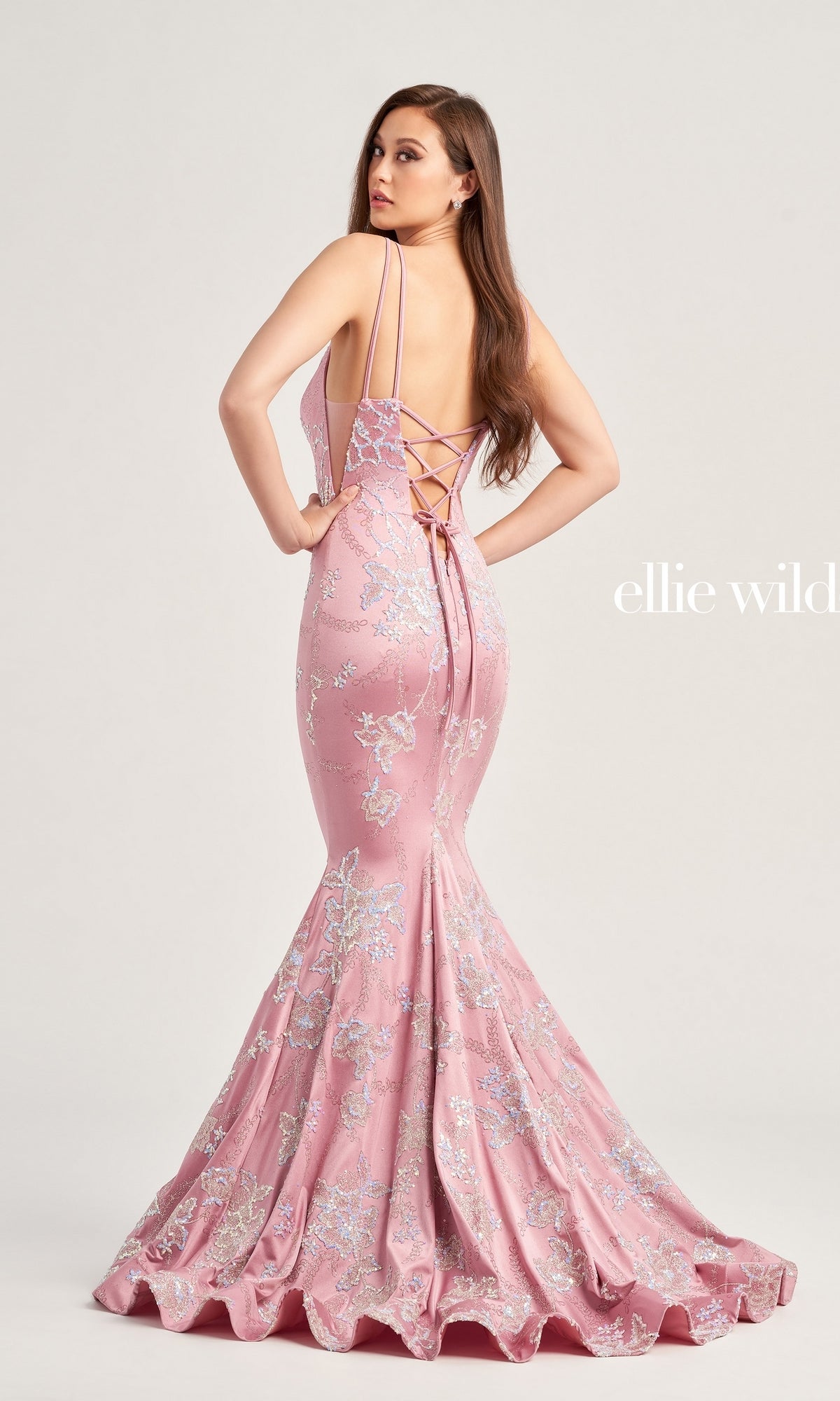 A woman in a pink Ellie Wilde Long Mermaid Formal Dress EW35083 with lace-up detailing and floral embellishments stands facing slightly away from the camera.